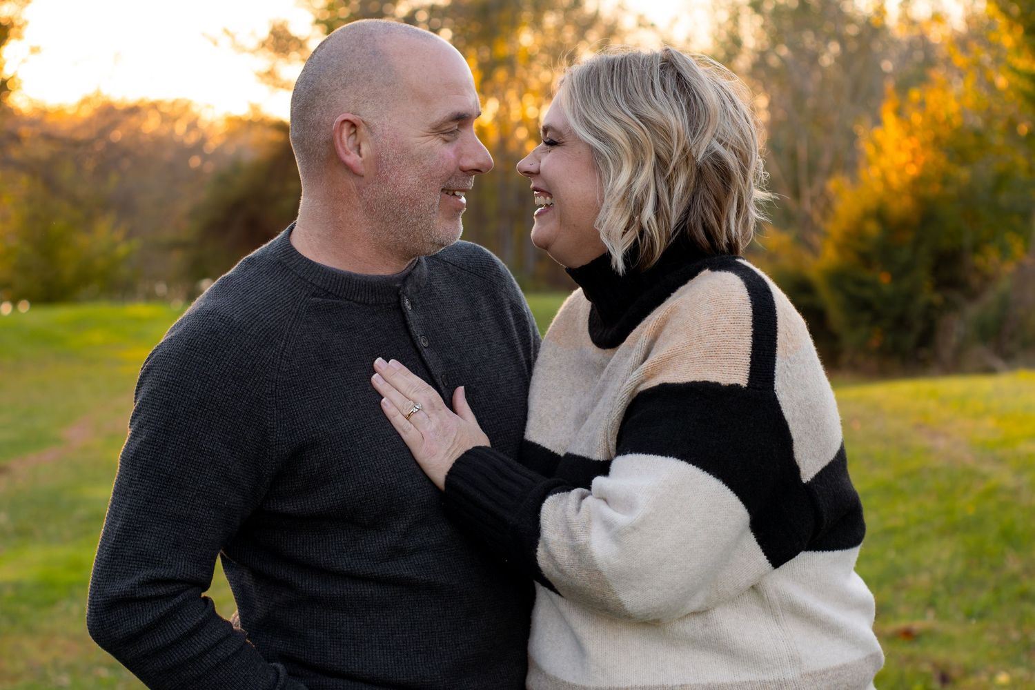Smiling couple embracing outdoors on a grassy field, the woman in a cream and black sweater resting her hand on the man's chest at sunset.