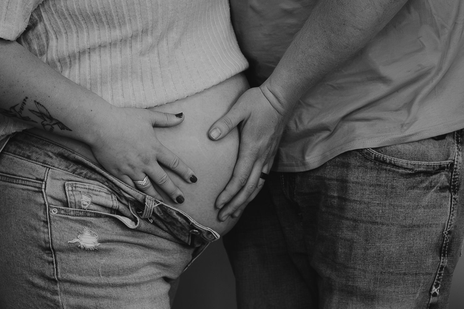 Black and white photo of a pregnant woman and partner tenderly holding her baby bump while wearing jeans.