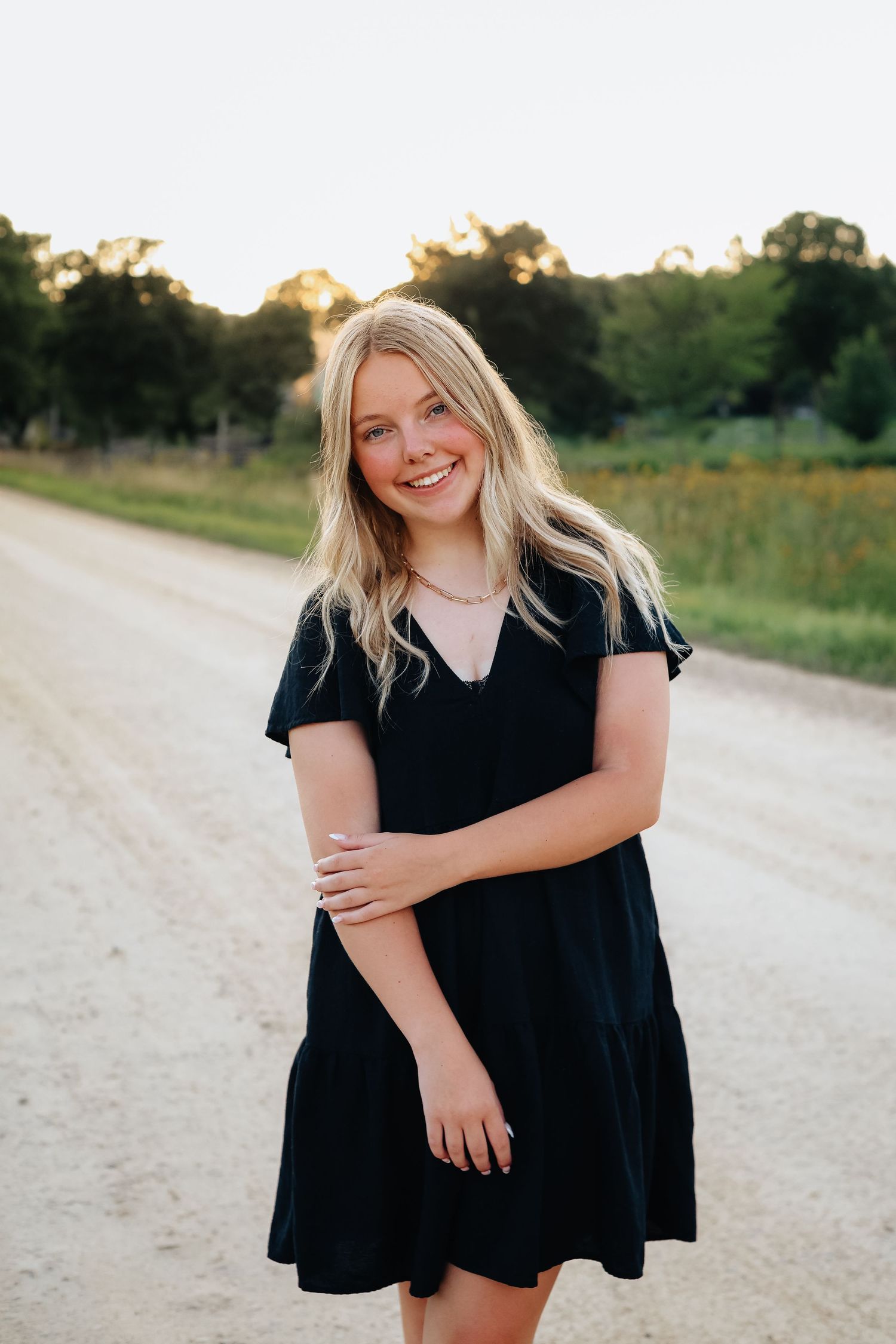 A woman in a black dress poses on a country road during golden hour.