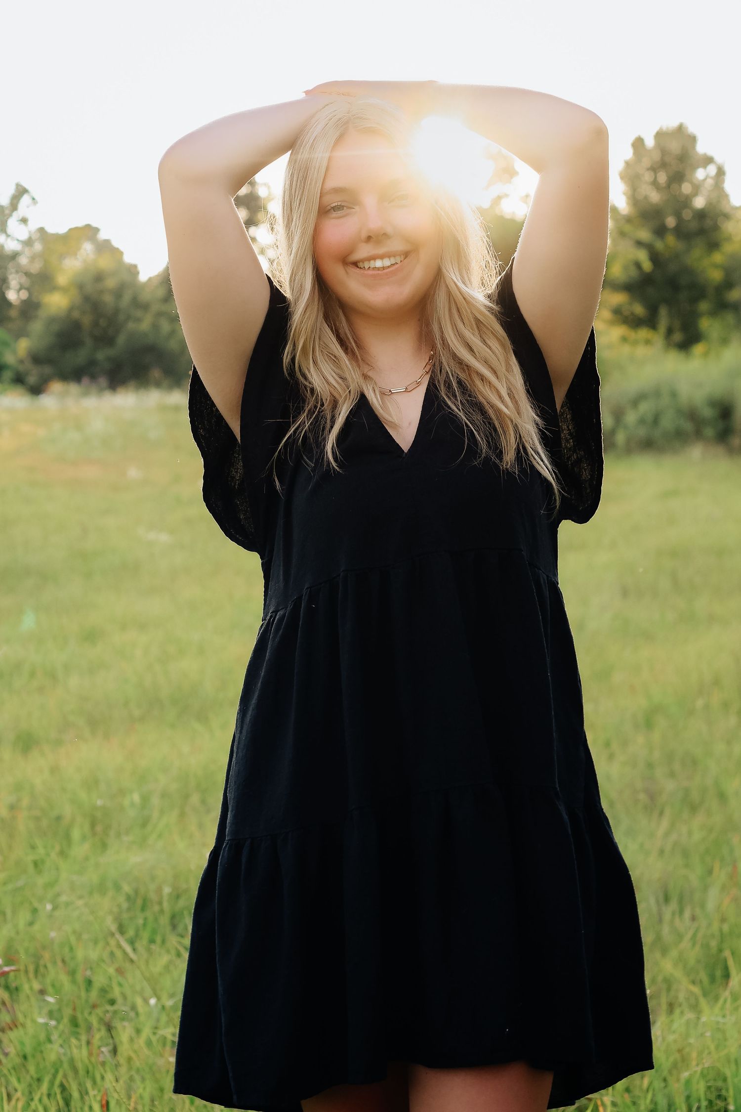 A woman in a black dress poses with arms raised during sunset in a field.