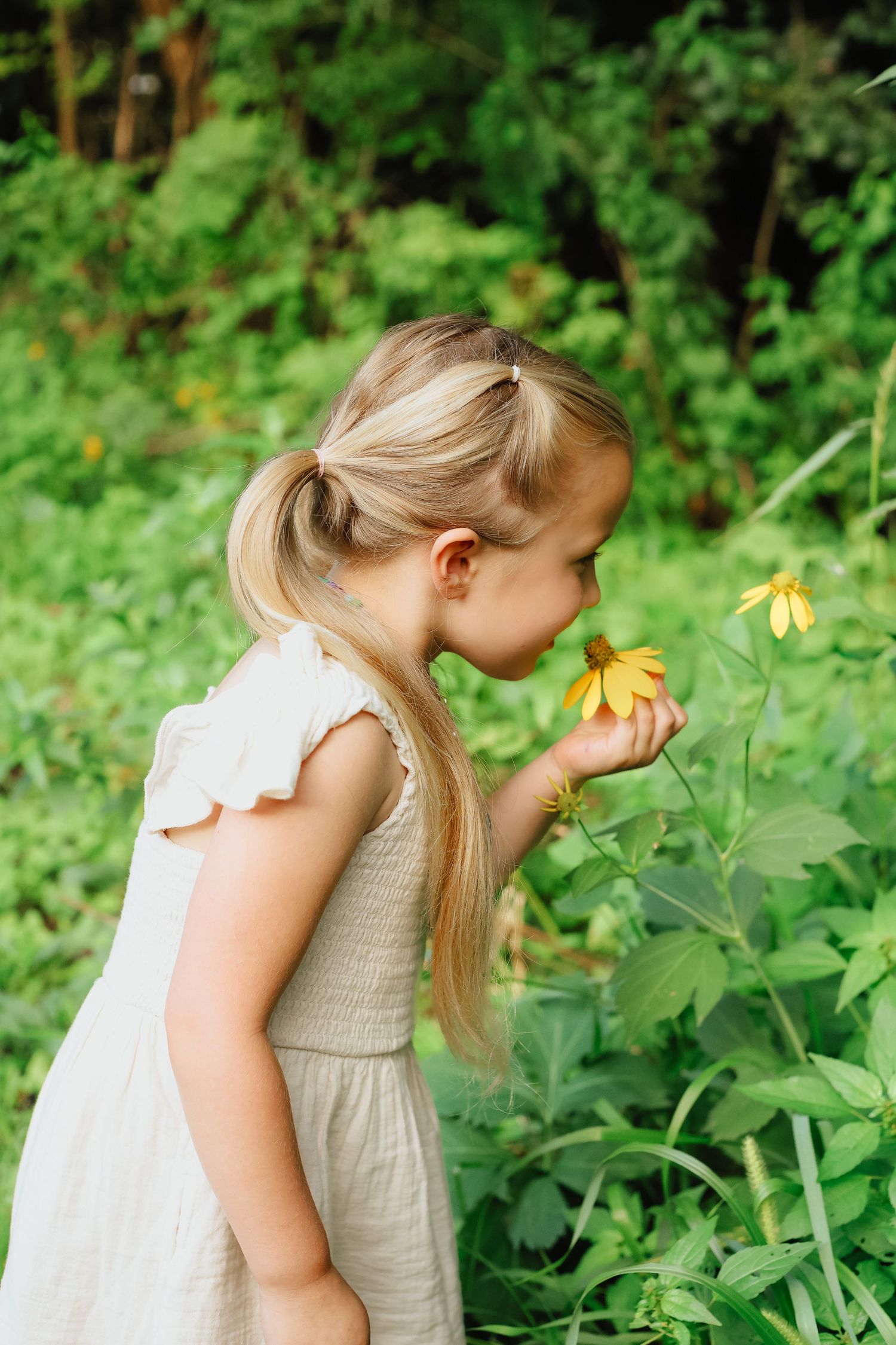 Child in white dress bending down to smell yellow wildflowers in a lush green garden.