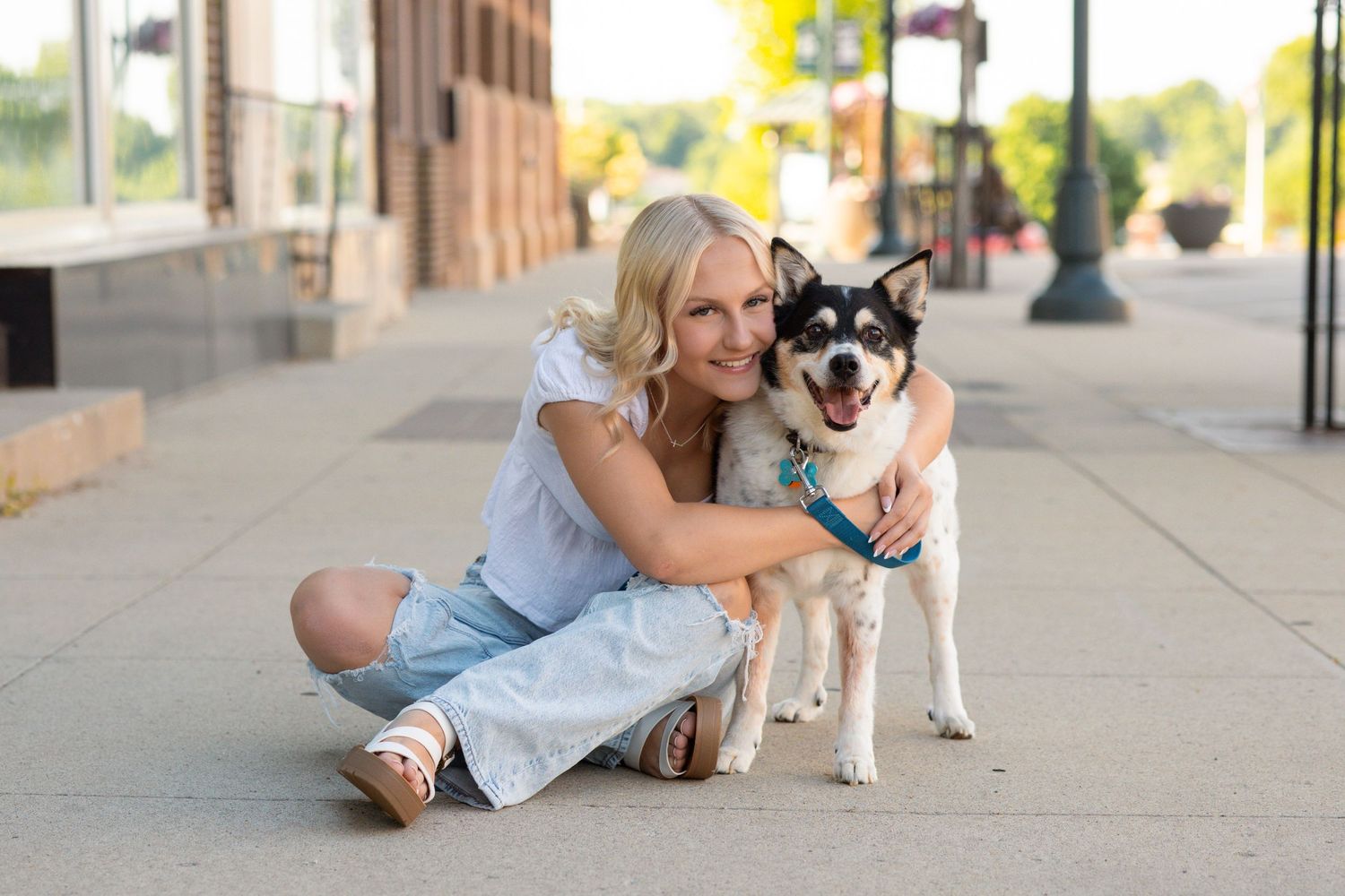 Smiling blonde woman sitting on sidewalk hugging a black and white Husky dog in an urban outdoor setting.