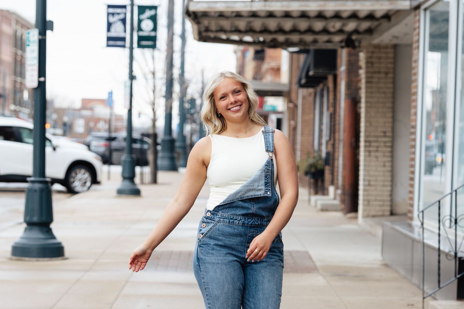 Young blonde woman smiling in denim overalls on a small-town sidewalk, posing confidently on a cloudy day.