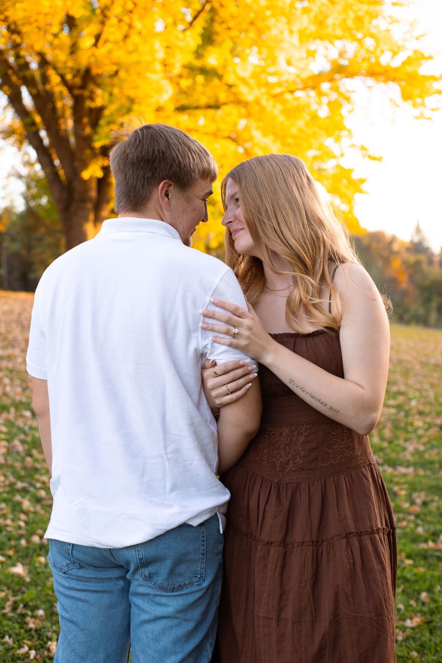 Couple embracing in a park beside a golden autumn tree, the woman in a brown sundress and the man in a white polo and jeans.