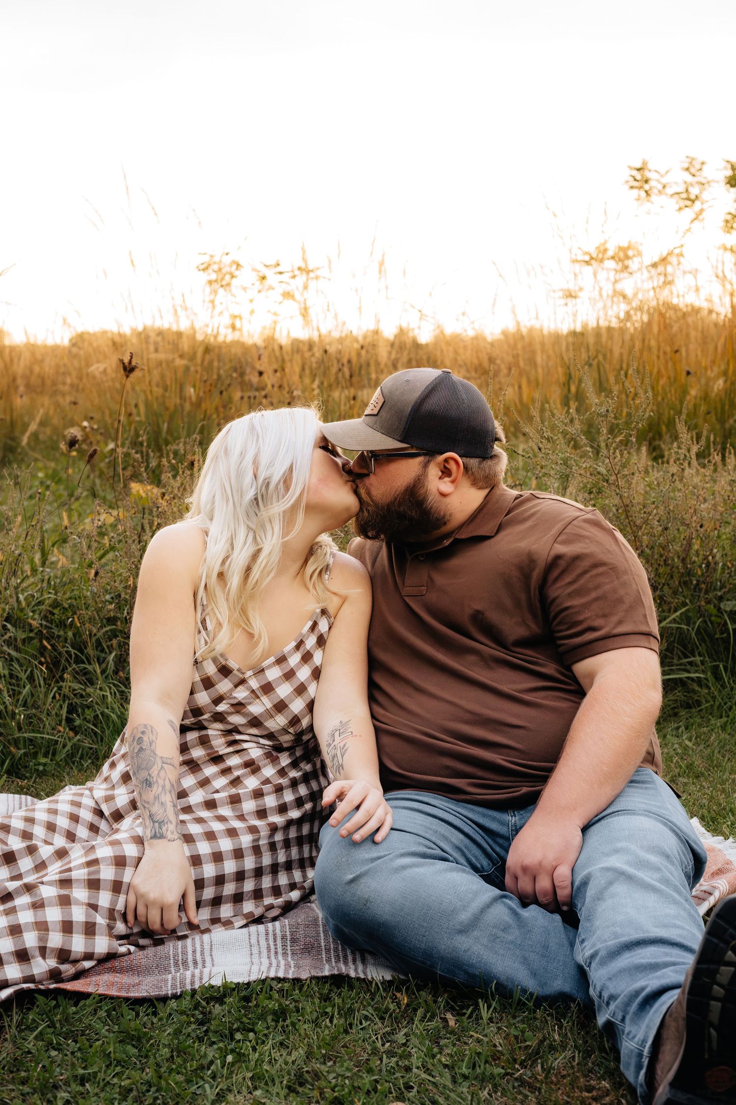 Couple kissing while seated on a plaid picnic blanket in a golden grassy field at sunset, wearing a checkered dress and a brown polo.