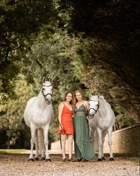 A group of white horses walk along a tree-lined path with people in orange and green dresses.