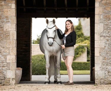 A person in shorts leads a white horse through a stone archway passage on a sunny day.