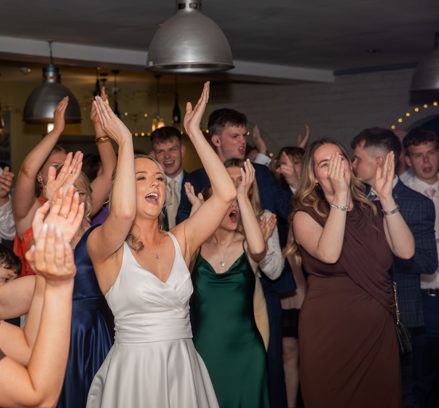 Excited wedding guests dancing and celebrating with hands raised at a lively indoor wedding reception party.