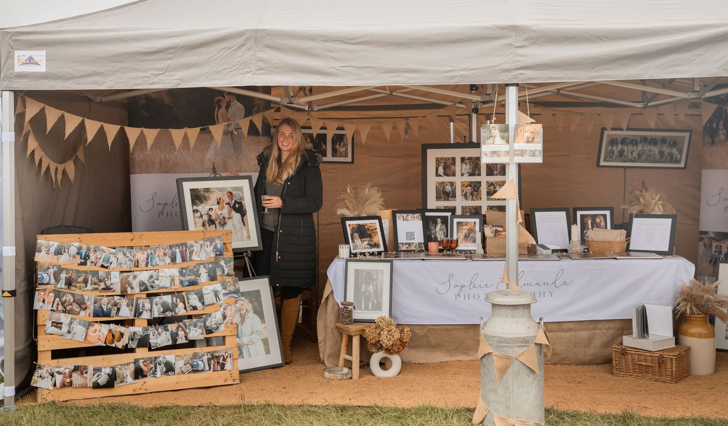 A vendor booth displays framed photo prints and collages under a tent at an outdoor market or craft fair.