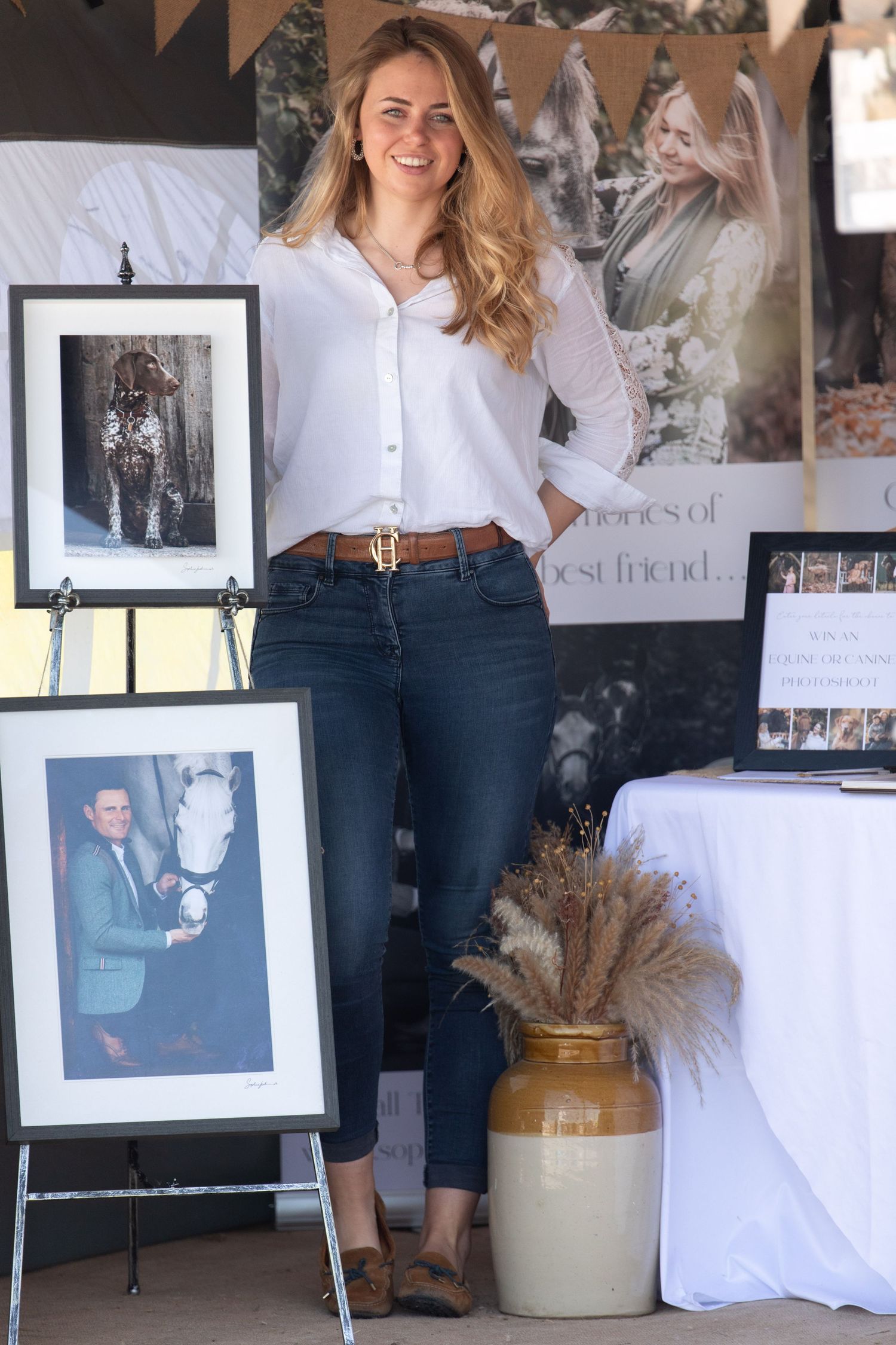 A photo studio setup with framed prints, dried flowers in a ceramic vase, and casual fashion styling against white walls.