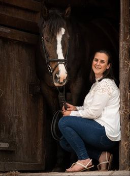 Woman in white blouse sits on wooden bench in rustic barn stall with horse visible in background.