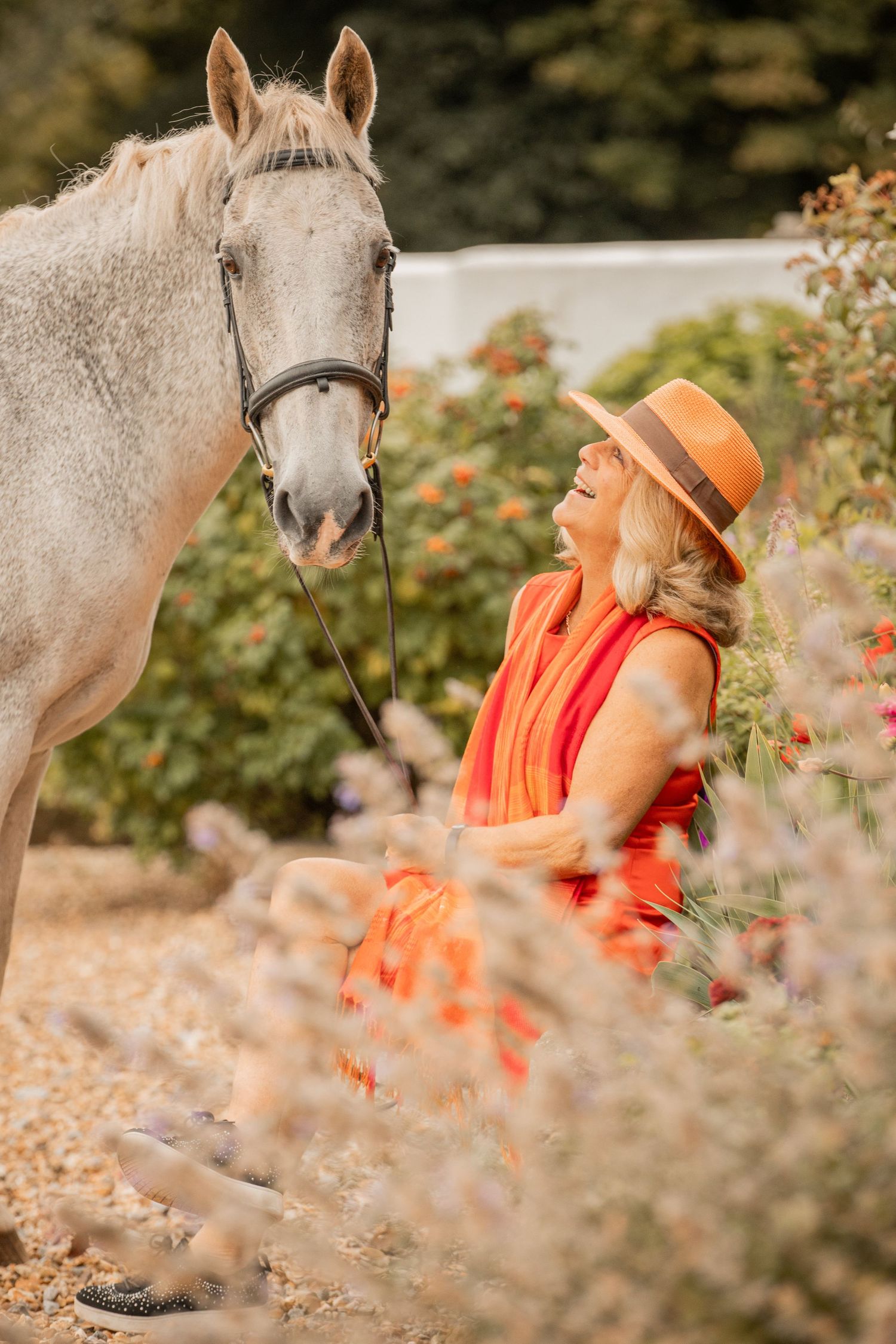 A white horse stands near orange flowers while someone in bright orange clothing sits in a meadow.