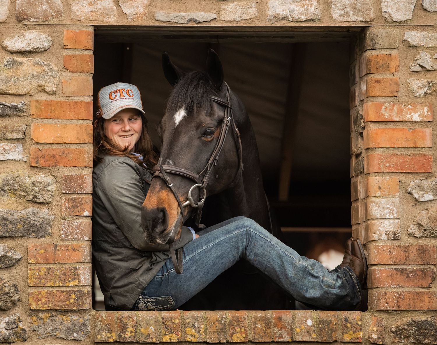 A person in casual attire sits in a rustic brick window frame with a beautiful black horse looking through the opening.