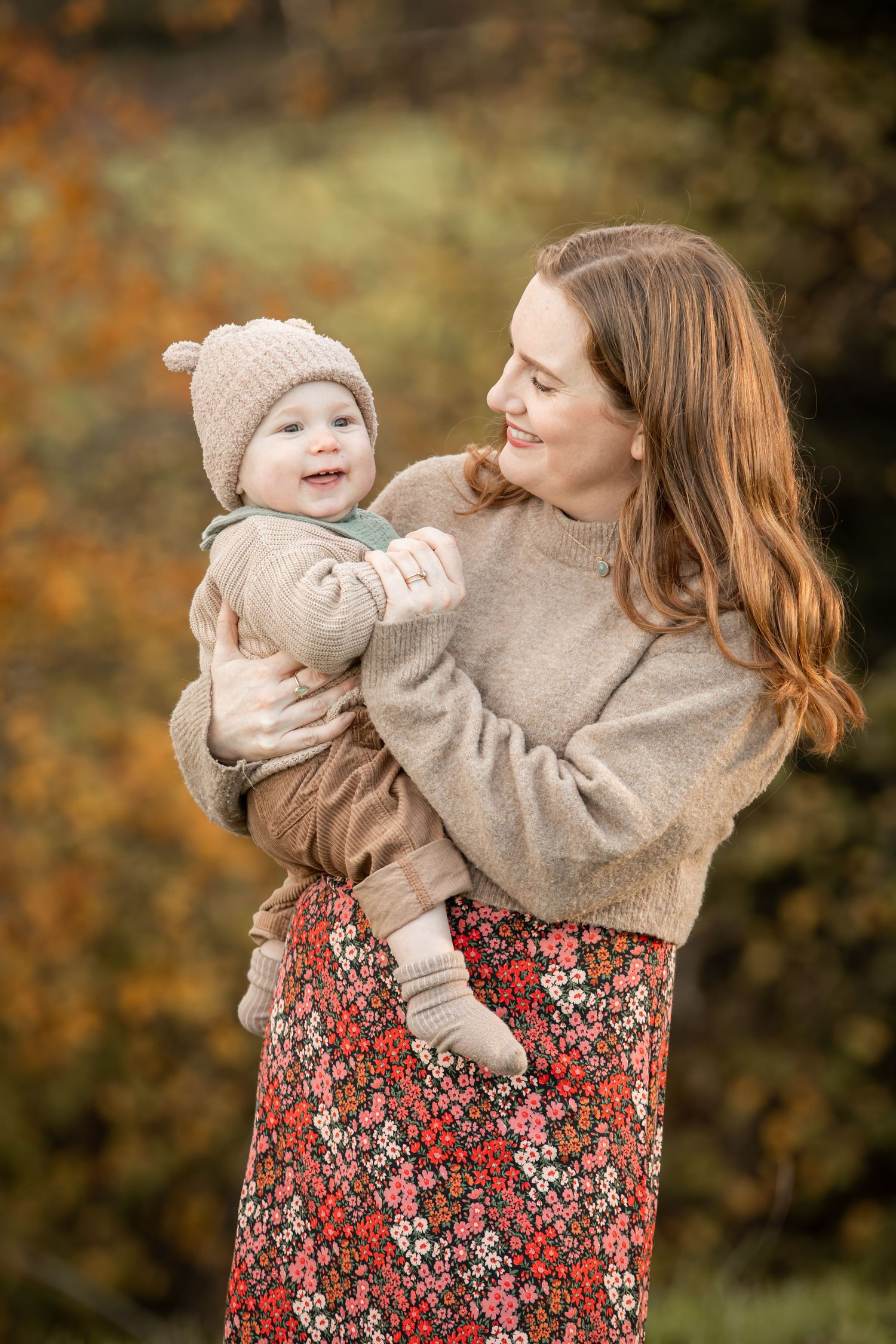 A parent and child in matching beige sweaters and floral dresses share joyful moments during an autumn photoshoot.