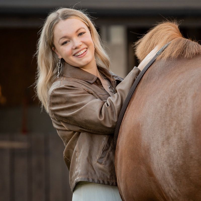 A person wearing a brown leather jacket smiles while standing next to a chestnut colored horse in a stable.