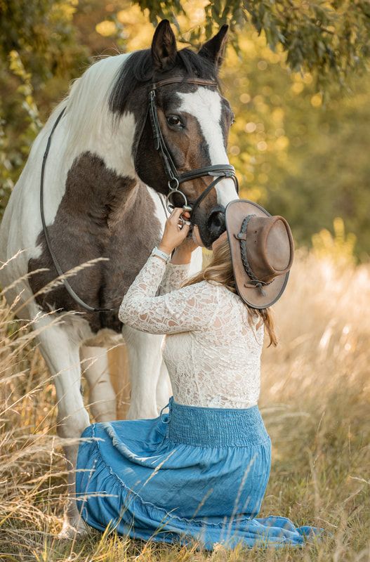 A grey and white horse wearing a bridle stands in a golden field with a person in a white lace top and blue skirt.