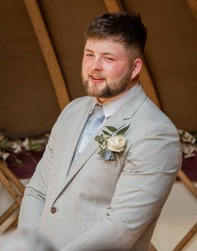 Groom in light gray suit with white boutonniere smiles during wedding ceremony in rustic tent setting.
