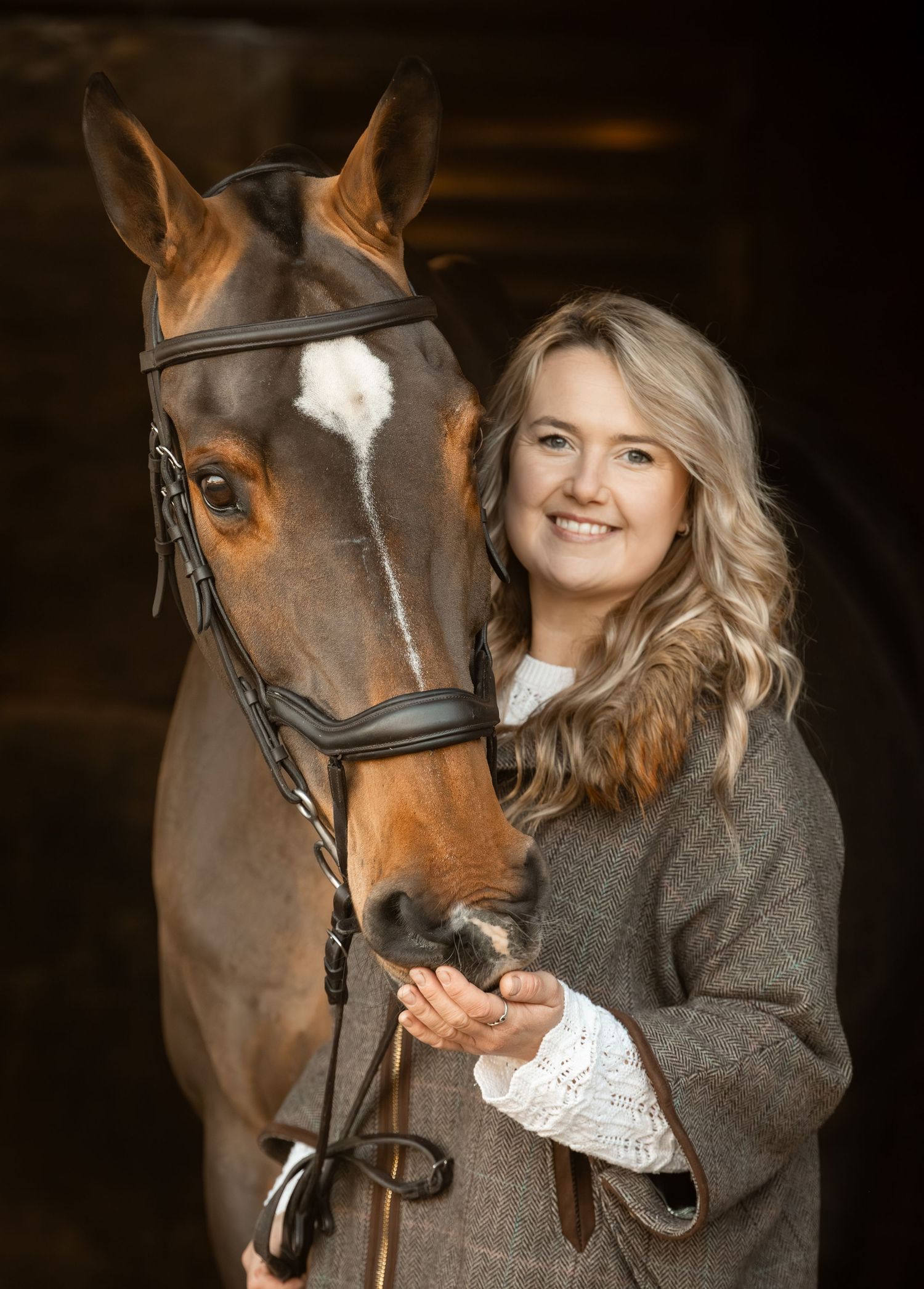 A bay horse with a white blaze stands with a companion in a dark barn wearing a black bridle and leather tack.