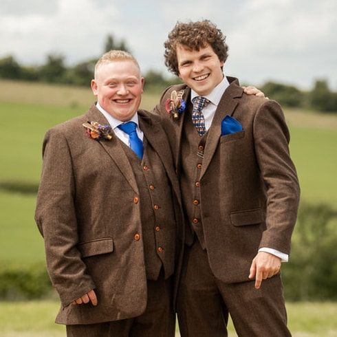 Wedding party members in brown tweed suits pose together on grassy field in countryside setting.