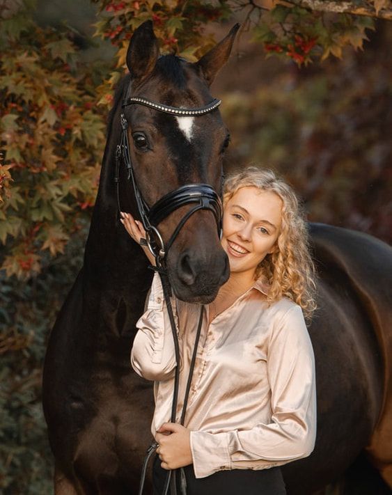 A black horse and rider in a pink blouse pose together against an autumn backdrop with red maple leaves.