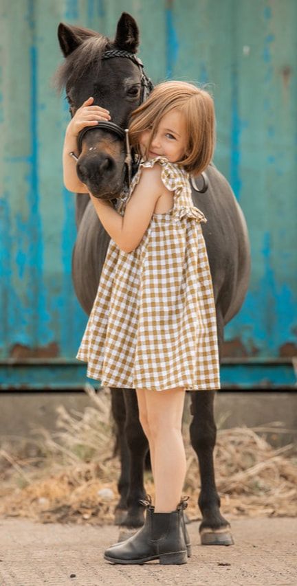 A young child in a brown checkered dress hugs a miniature black pony against a blue wall backdrop.