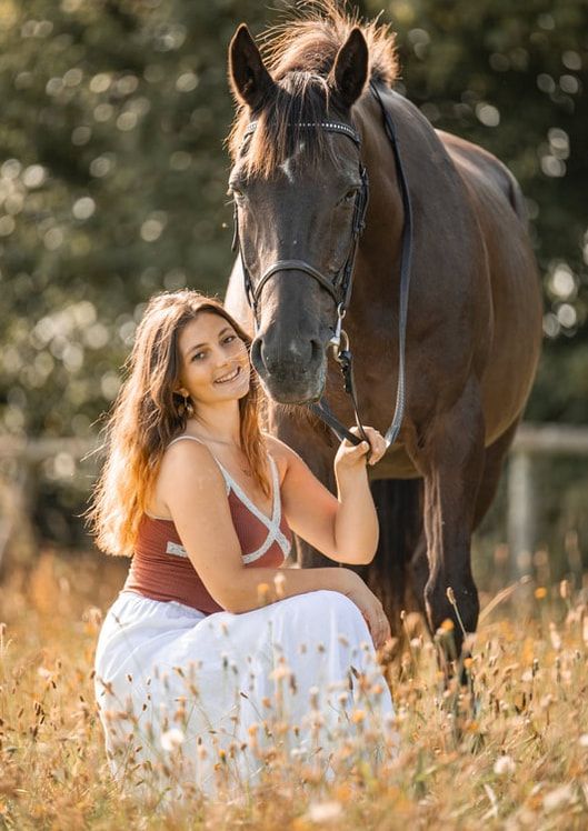 A person in a white dress sits in golden grass beside a dark brown horse during a sunset photoshoot.