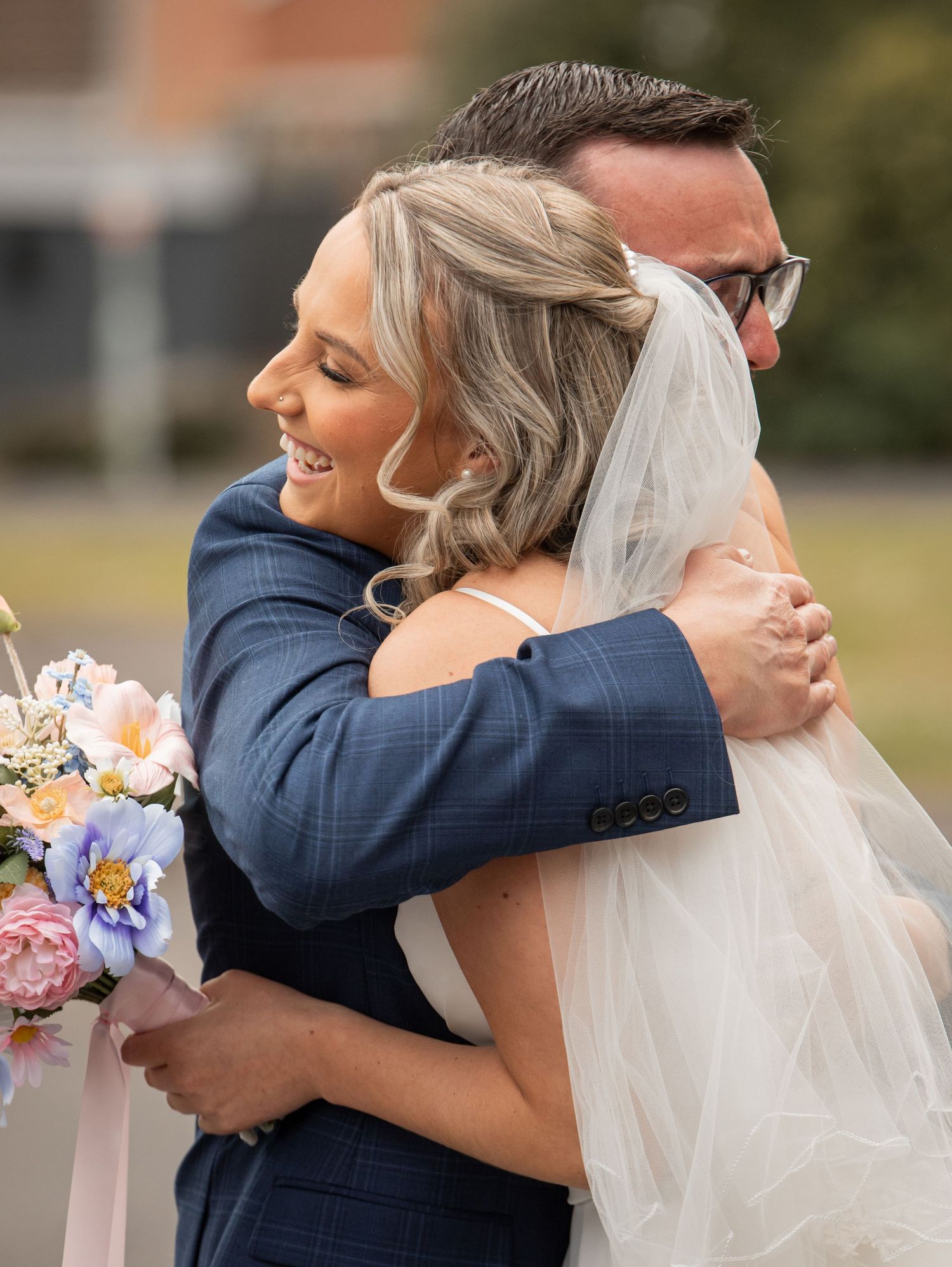 A bride in a white gown and veil embraces a groom in a blue suit, smiling joyfully while holding a pastel bouquet.