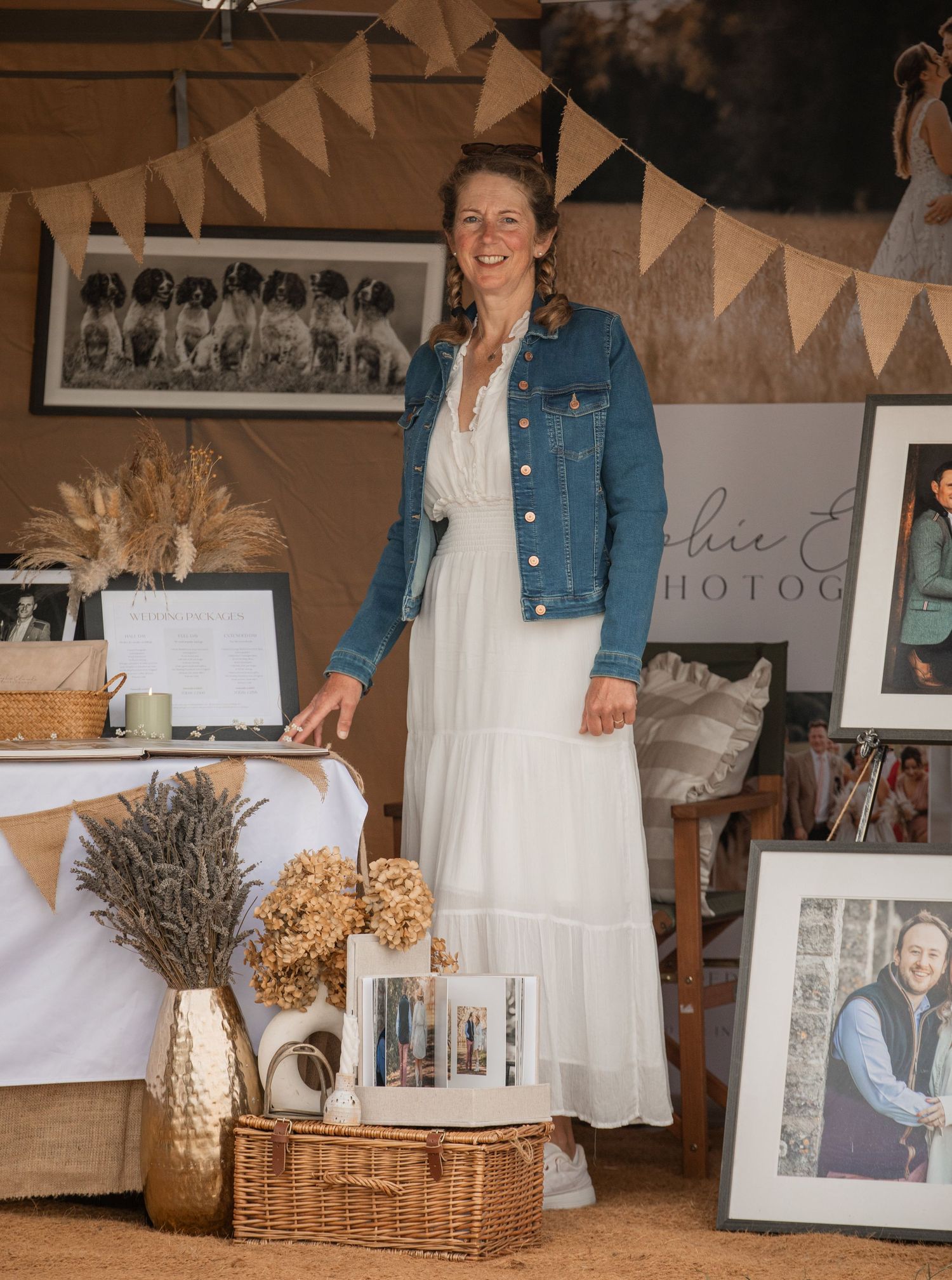 Woman in white dress and denim jacket stands in a rustic photography display booth with framed prints and dried flowers.