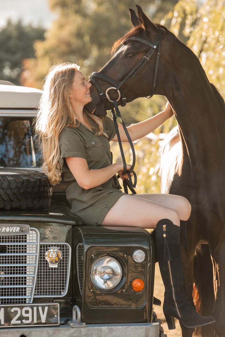 A woman in olive green sits with a black horse beside a vintage Land Rover during golden hour lighting.