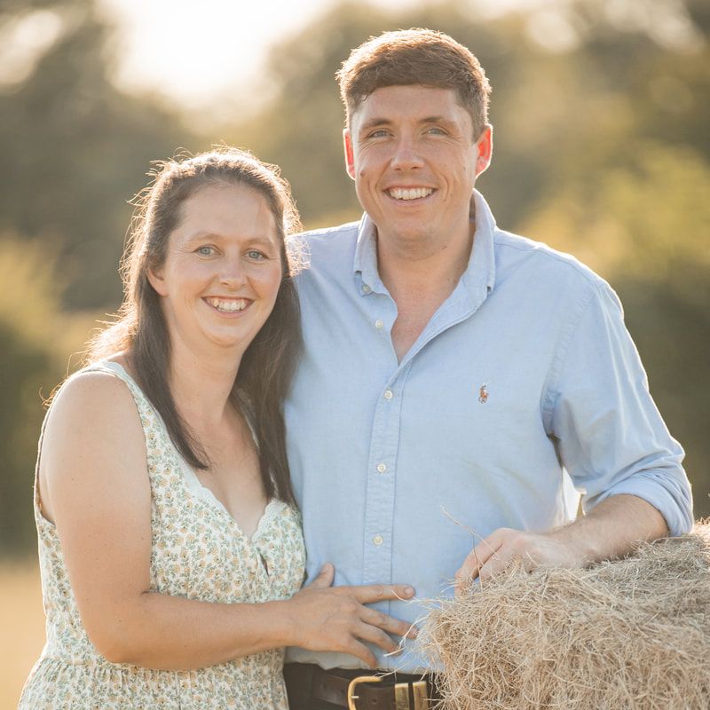 A couple poses together on hay bales during a golden sunset photoshoot in a rustic outdoor setting.