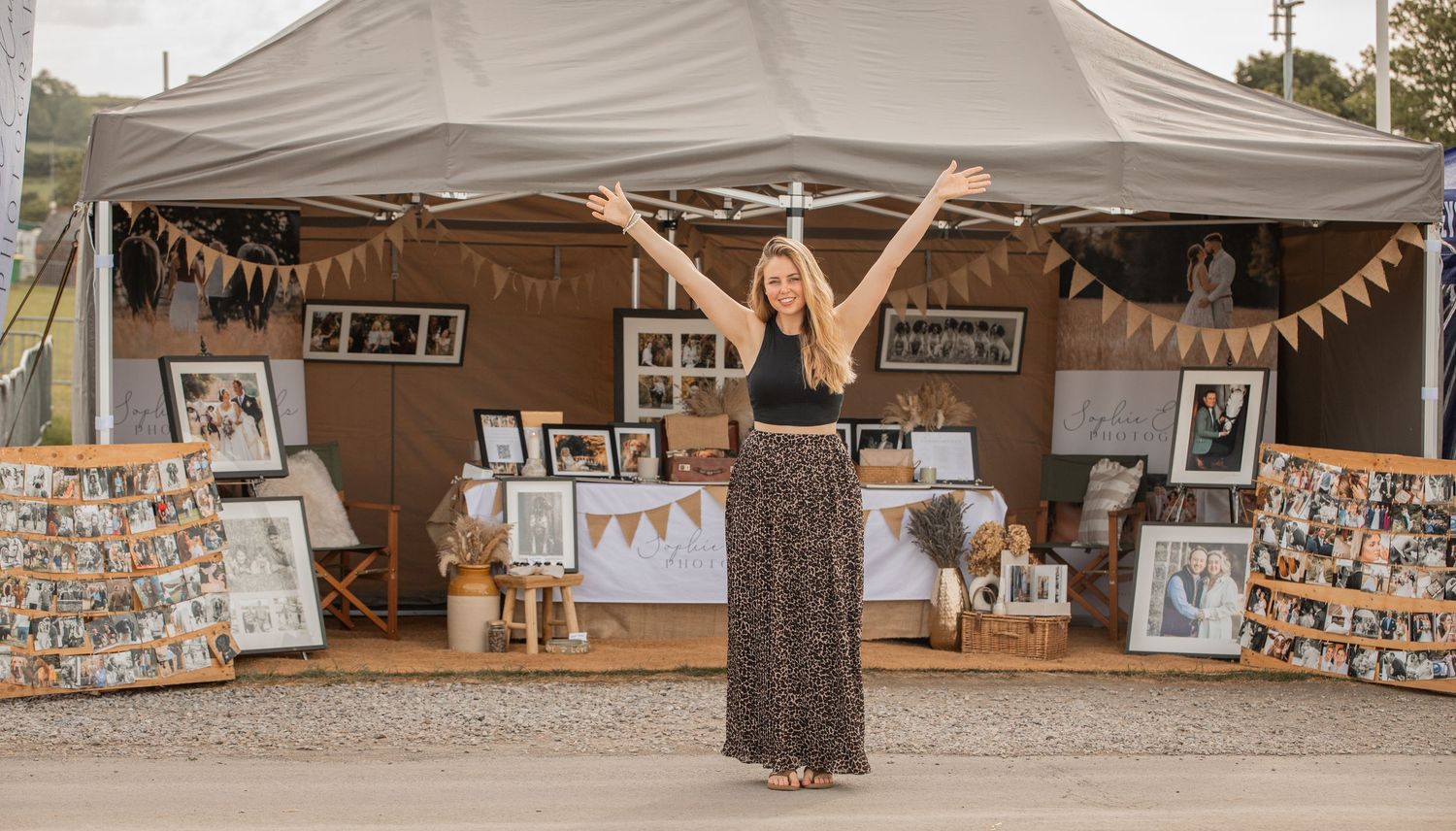 Woman with arms raised stands before an outdoor photography display booth with framed portraits and bunting decorations.