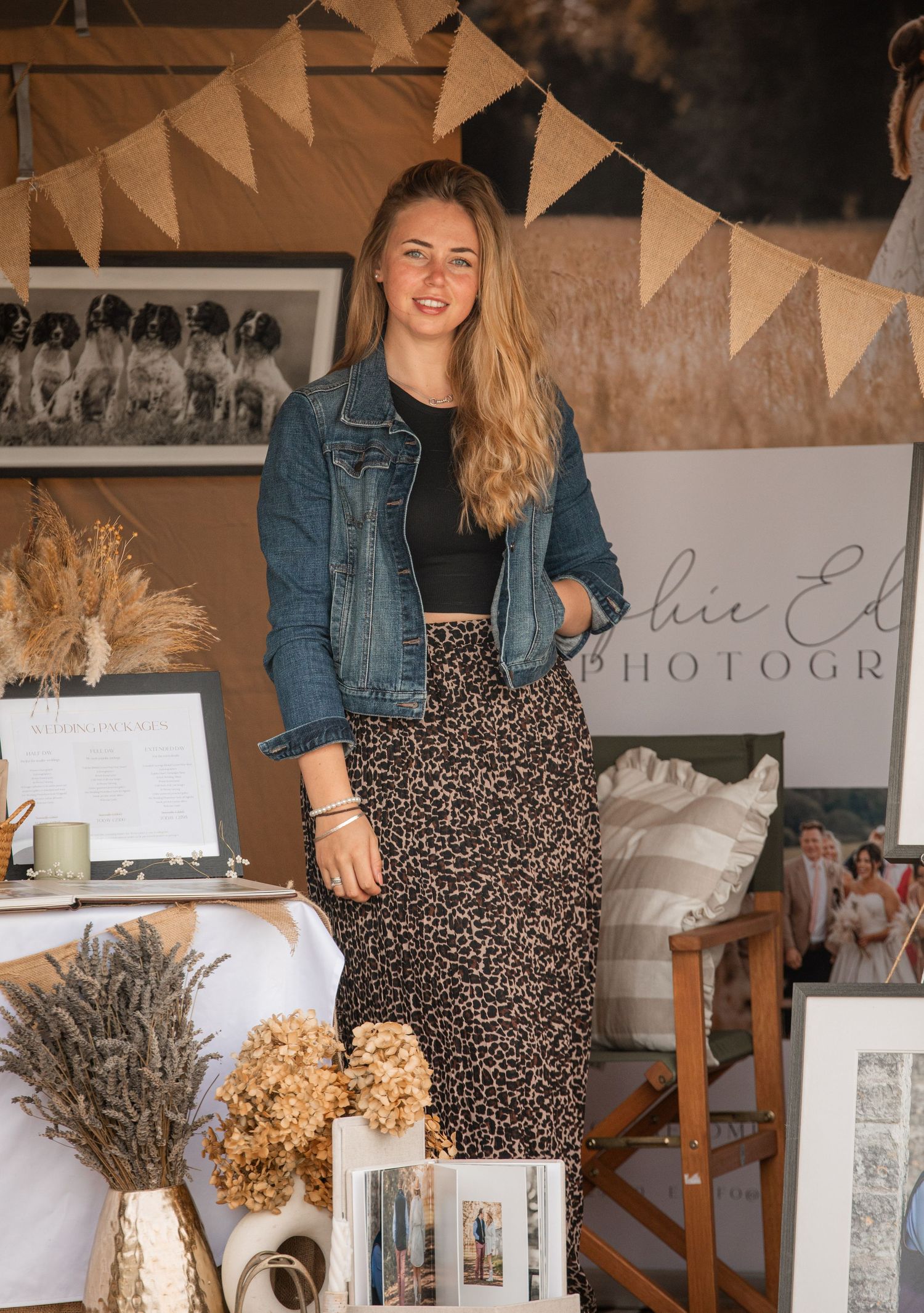 Young woman in denim jacket and leopard print skirt stands smiling at a photography business display booth.