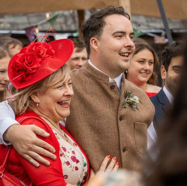 A group of people at an outdoor event wearing formal attire including a bright red hat and brown coats.