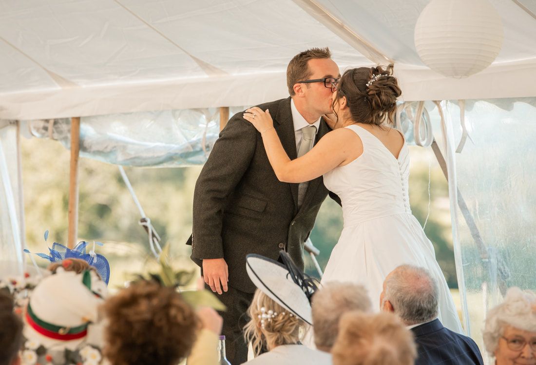 Couple shares their first kiss as newlyweds under a white tent during an outdoor wedding reception celebration.
