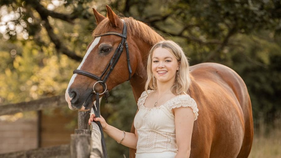 A woman in a white lace dress poses with a chestnut horse in a sunlit outdoor setting during a photo session.