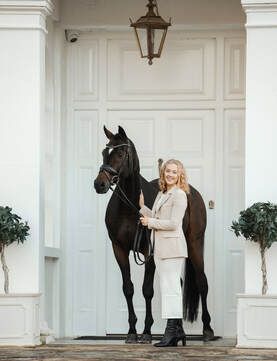 A woman in riding attire stands with a majestic black horse against a white columned building exterior.