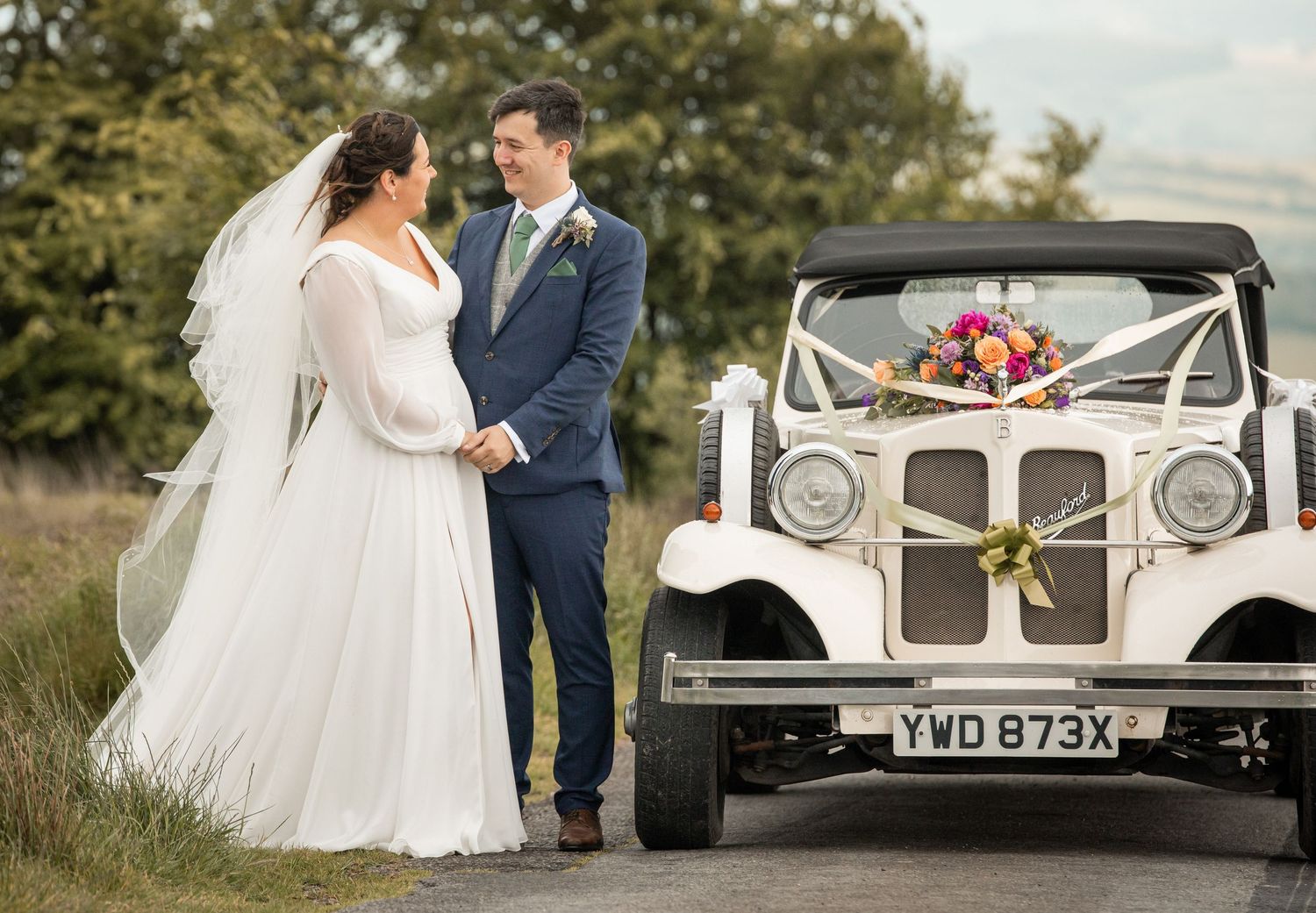 Romantic wedding couple poses next to a classic white vintage car decorated with floral arrangements in a countryside setting.