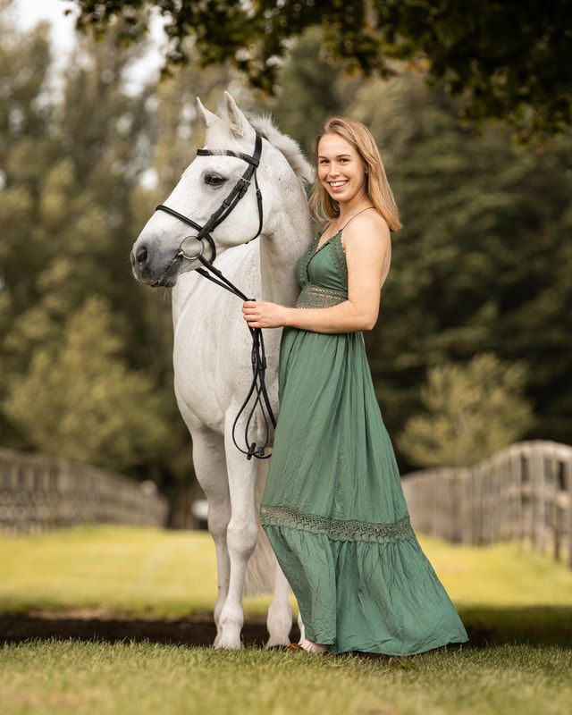 A person in a green dress stands beside a majestic white horse in a sunny outdoor field setting.