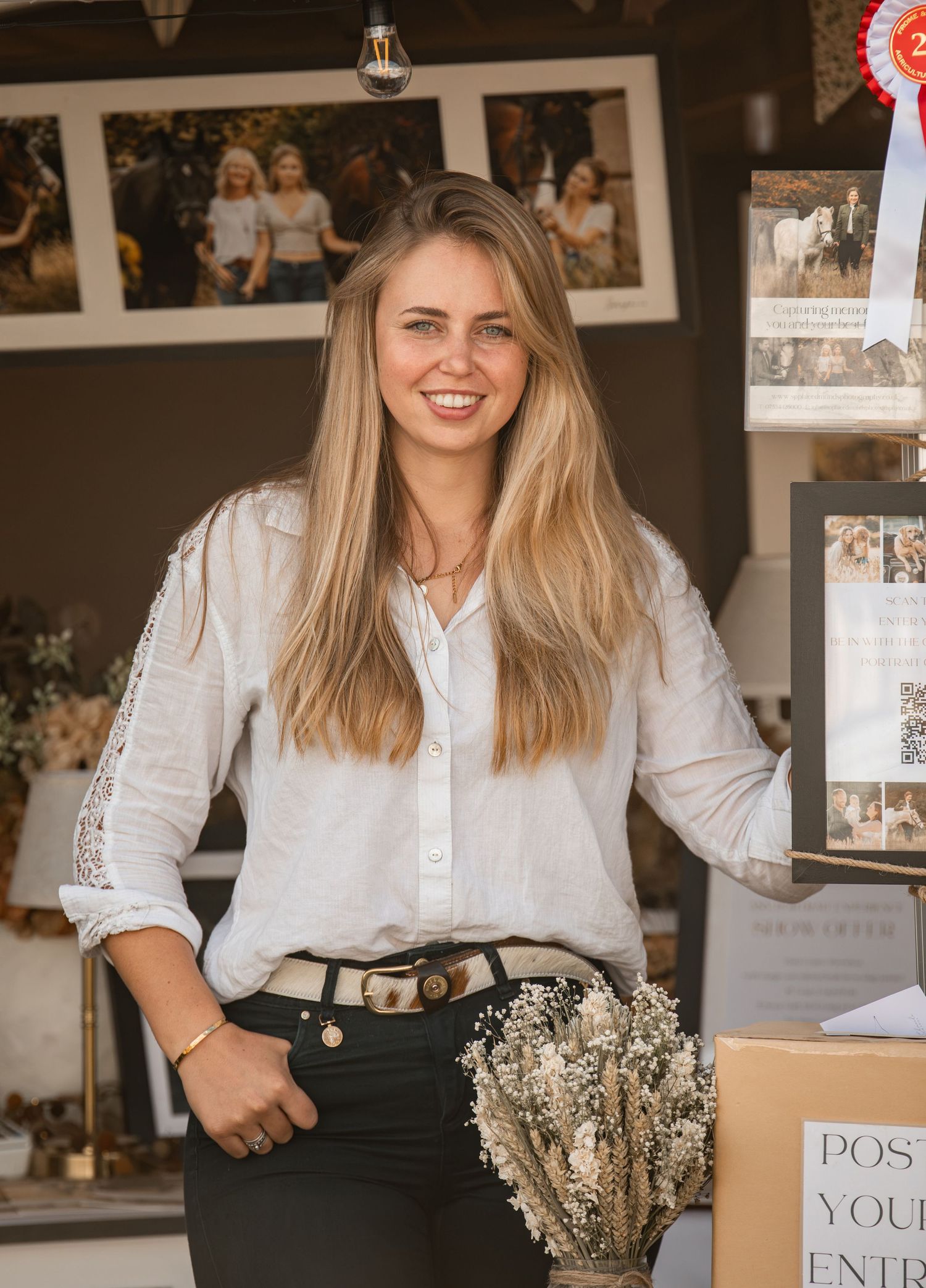 Smiling blonde woman in white lace blouse holding dried flowers at a market stall display.