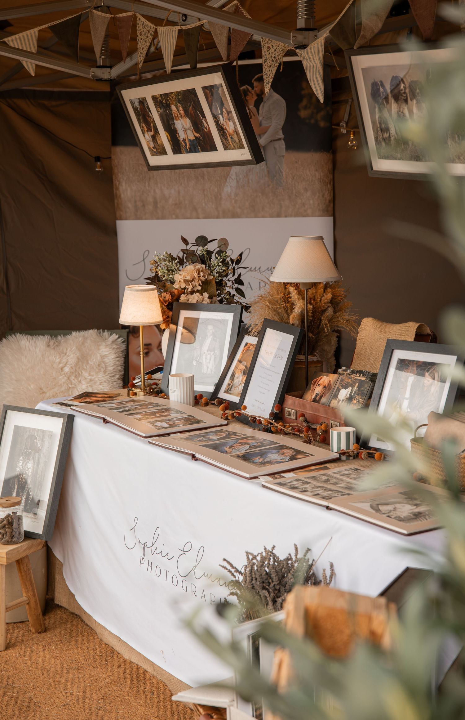 Elegant photography booth display with framed prints, floral arrangements, and albums on a white draped table at a wedding fair.