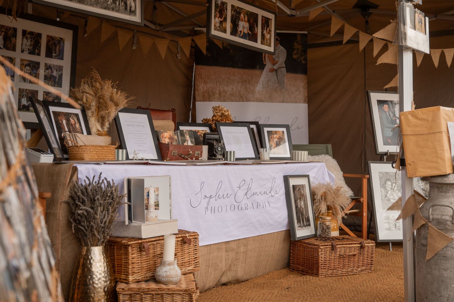 Rustic wedding display with framed photos, wicker baskets, dried flowers, and elegant calligraphy signage in a warm tent setting.