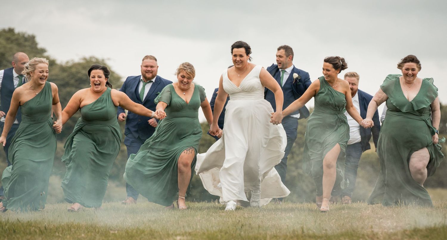 Wedding party members in green dresses and formal attire jump joyfully together in an outdoor field setting.