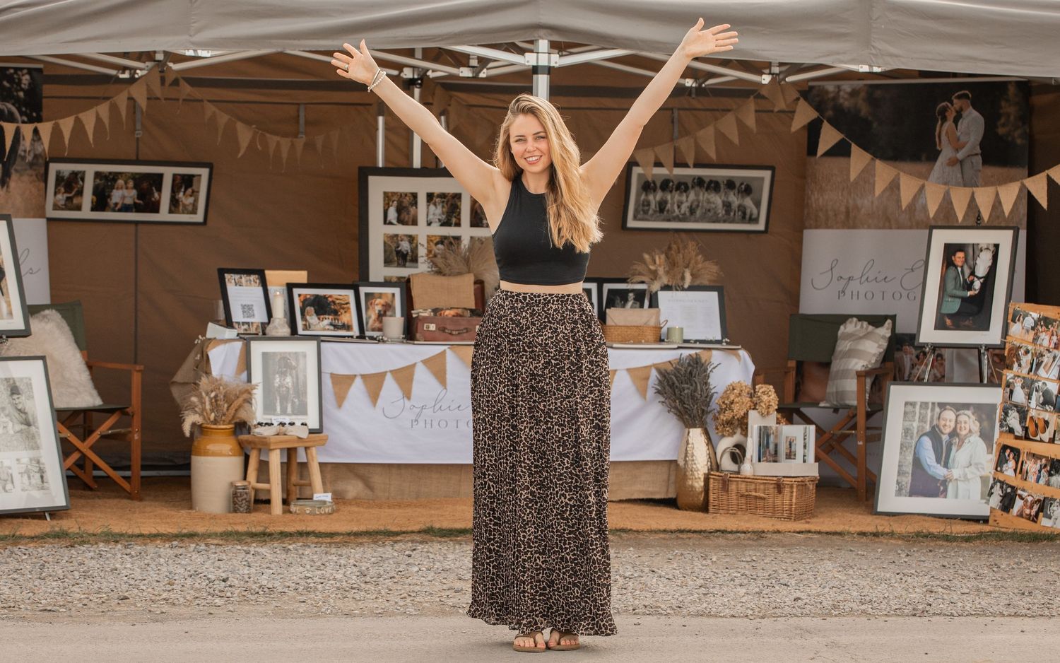 Woman with arms raised at a photography booth displaying framed portraits and wedding photos at an outdoor market.