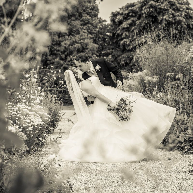 A couple in wedding attire embraces while lying in a grassy outdoor setting, creating a romantic black and white portrait.