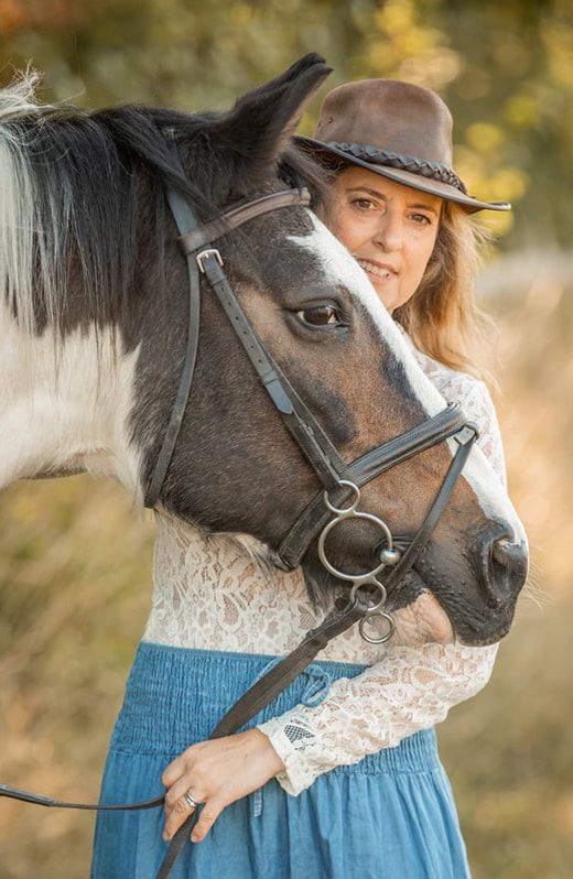 A black and white horse wearing a bridle stands next to someone in a cowboy hat, blue skirt and lace top at sunset.