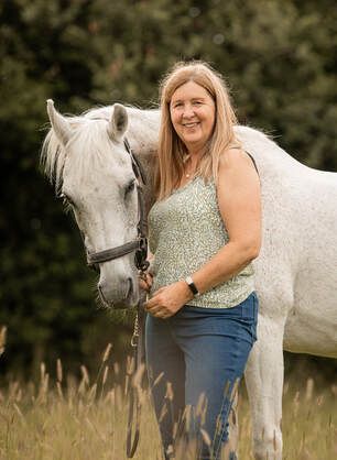 A woman in a floral top and jeans poses with a beautiful white horse in a grassy outdoor setting.