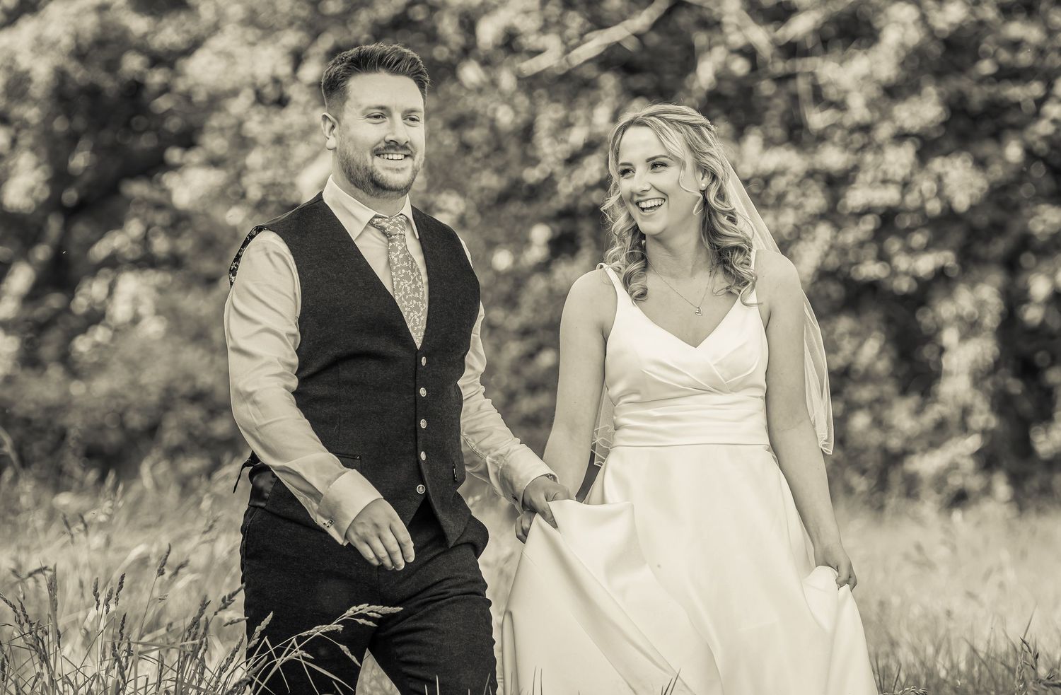 Black and white photo of a smiling bride and groom walking hand in hand outdoors on their wedding day.