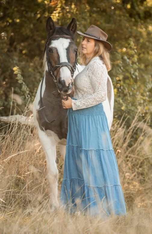 A person in a blue flowing skirt and lace top stands with a black and white horse in a sunlit meadow.