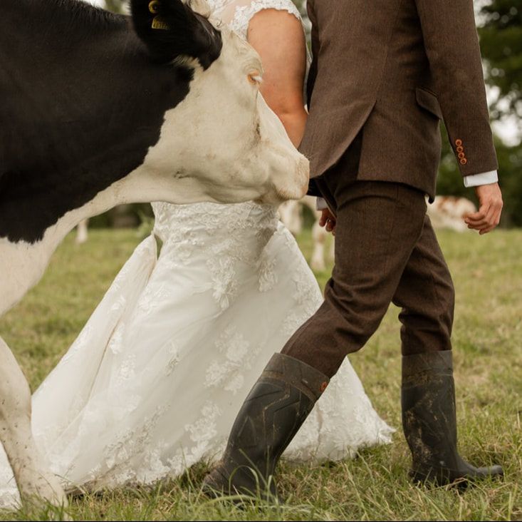 Close-up of bride in white lace dress and groom in brown suit wearing boots standing on grassy field.