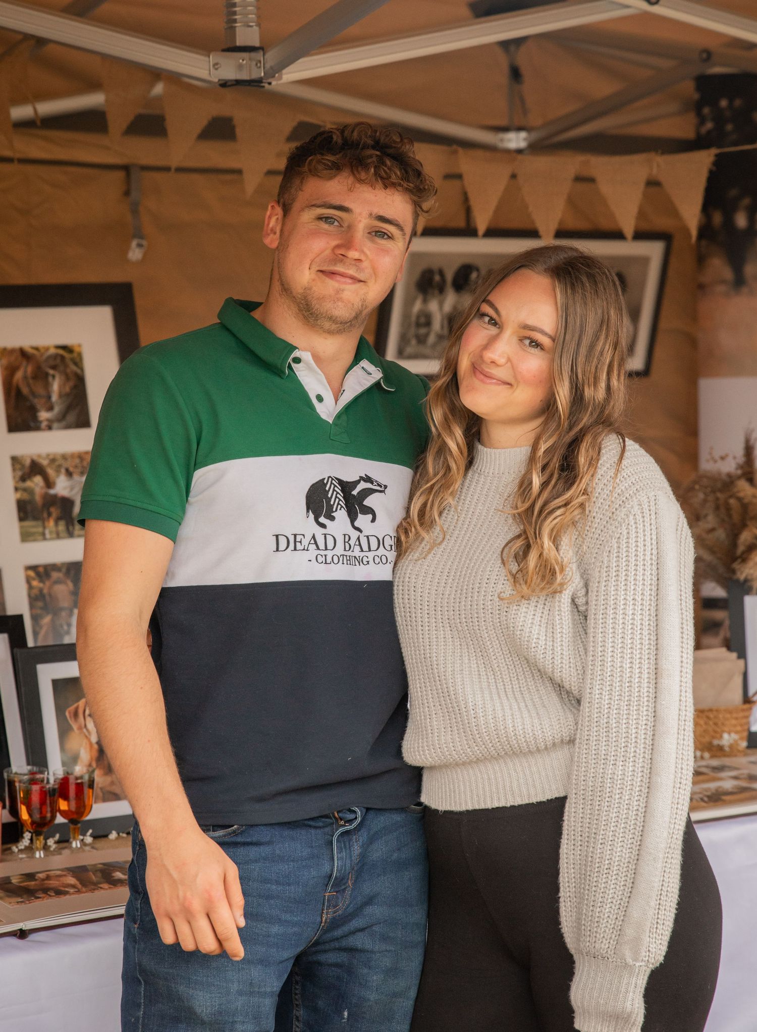 A young couple posing together at an outdoor market stall, man wearing a Dead Badger polo shirt, woman in a grey knit sweater.